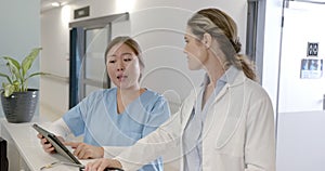 Focused diverse female doctors using tablet and discussing at hospital reception desk, slow motion