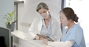 Focused diverse female doctors using tablet and discussing at hospital reception desk, slow motion