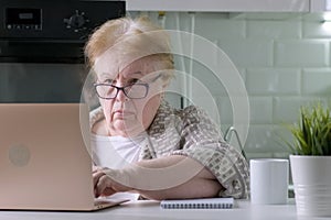 Focused elderly woman sitting at the table at home working using computer laptop with serious face