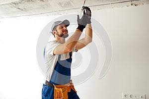 Focused electrician in uniform installing light fitting in new apartment, standing on a ladder