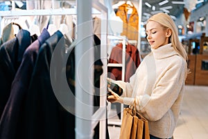 Focused elegant blonde young woman choosing coat in clothing store, standing near showcase with female clothing, sorting