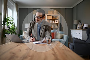 Focused elegant businessman signing documents while working over laptop on desk in home office
