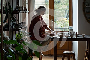 Focused enterprising woman ceramist sits at table with white crockery in workshop of pottery shop