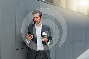 Focused european businessman in suit with phone drinking coffee near office building during break