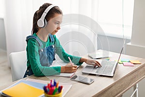 Focused girl sitting at desk, using laptop, writing in copybook