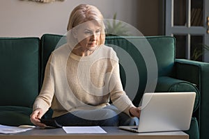 Focused grey haired mature woman calculating bills, using laptop