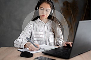 Focused hispanic woman wearing headphones using laptop at home, writing notes, female college student learning and