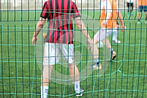 Focused image of sectional green fence. Soccer players with a ball plays on the background