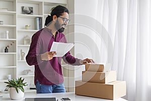 Focused Indian businessman reviewing documents and organizing shipping boxes at home office