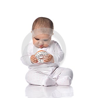 Focused infant baby toddler in white onepiece overall sits on the floor occupied with donut toy in hands