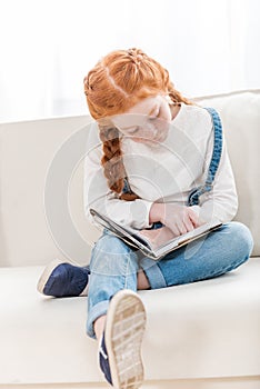 Focused little girl reading book while sitting on sofa at home