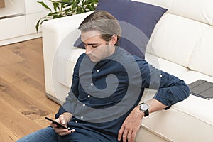 A focused looking handsome young man sitting on the ground and leaning on a sofa checking his phone