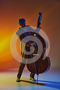 Focused male African-American artist in coat, performing on upright bass, in warm neon light against gradient background