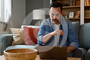 Focused male computer programmer holding eyeglasses and working over laptop seriously while sitting on sofa. Young pensive