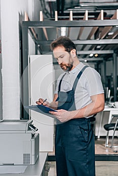 Focused male craftsman checking copy machine and writing in clipboard photo