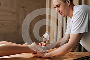 Focused male masseur massaging feet using herbal bolus bags to young woman lying on massage table. Professional