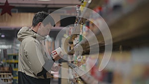 Focused man reads labels on products in supermarket, carefully examines ingredients and expiration dates when buying