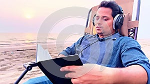 focused Man relaxing on beach recliner chair reading a book while listening music on headphone by the sea