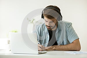 Focused man wearing headphones writing notes learning online wit