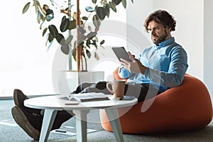 Focused mature entrepreneur using digital tablet at workplace, sitting in bean bag chair at office, copy space