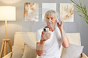 Focused mature man attired in white T-shirt sitting on sofa holding his mobile phone and headphones enjoying online music photo