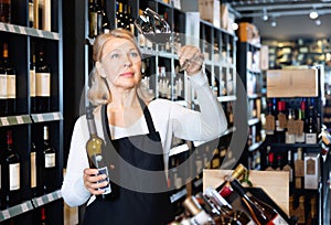 Focused mature woman wine producer inspecting quality of wine in wineshop on background with shelves of wine bottles