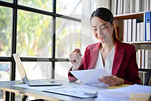 Focused millennial Asian businesswoman examining financial report, working at her desk in office