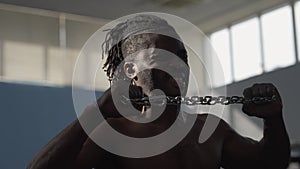 Focused motivated sportsman pulling metal chain with hands indoors. Perspiring handsome African American man training