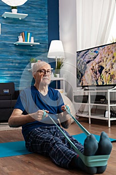 Focused pensioner sitting on yoga mat stretching legs muscle using fitness elastic band