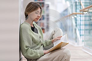 Focused pensive middle-aged woman passionate reader reading book while sitting on floor in library