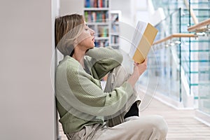 Focused pensive middle-aged woman passionate reader reading book while sitting on floor in library