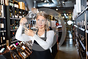 Focused mature woman wine producer inspecting quality of wine in wineshop on background with shelves of wine bottles