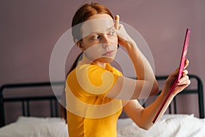 Focused redhead young woman with natural beauty applying powder or blusher on face with big makeup brush sitting on bed