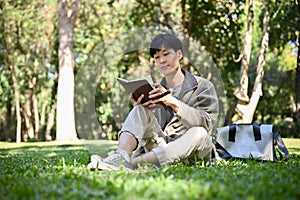 Focused and relaxed young Asian male college student chilling in the park, reading a book or novel