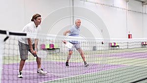 Focused resolved adult man playing friendly pickleball match in team with older partner on closed court. Concept of