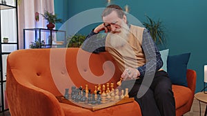 Focused senior grandfather man playing chess leisure board game alone, domestic activity at home