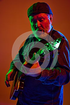 Focused senior guitarist in cap tuning guitar in colorful neon light against orange studio background.