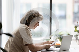 Focused senior homeowner, landlady reading paper document at laptop