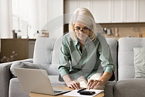 Focused senior retired lady in glasses typing on calculator