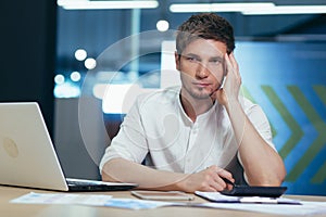 Focused and serious young man IT specialist, manager, businessman working on laptop and with documents in the office at the desk.