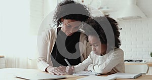 Focused small afro american girl doing homework with smiling mommy.