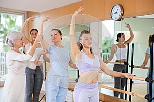 Focused sporty young women standing near ballet barre training positions with senior female coach during group lesson in