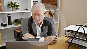 Focused and successful senior man boss sitting at the office desk, arms crossed in confident gesture, seriously looking at laptop