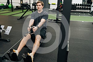 Focused successful sportsman working out on stationary rowing machine in gym. Young beginner sporty man doing exercises