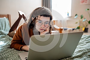 Focused teen girl studying, watching online lecture, webinar, seminar at laptop lying in bed at home