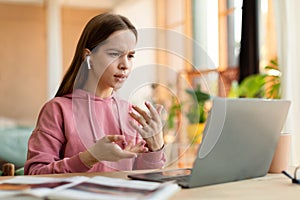 Focused teenage girl video calling, wearing wireless earphones and gesturing at laptop webcamera, sitting at home