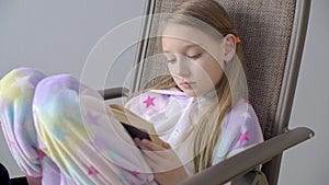 Focused teenager girl in pajama reading book in chair. Young schoolgirl in home clothes reading book sitting in chair in