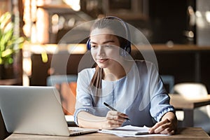 Focused woman wearing headphones using laptop, writing notes