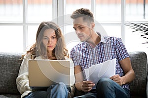 Focused worried couple paying bills online on laptop with document