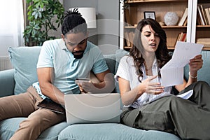 Focused worried couple paying bills online on laptop with documents sitting together on sofa at home, serious confused man and
